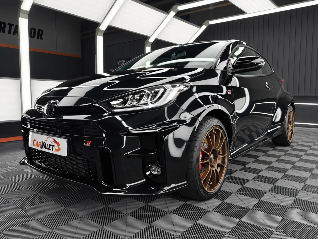 Schwarzer Toyota GR Yaris mit bronzefarbenen Felgen, schräg von vorn im Lichttunnel von Car and Valet.